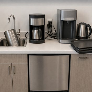 A breakroom counter run where a single wall outlet feeds three appliances through an adapter, the cords running loose across the work surface between a coffee brewer, a water dispenser and a kettle; at the end of the run a large pitcher stands tilted in a sink too shallow to hold it flat, its rim higher than the faucet spout.