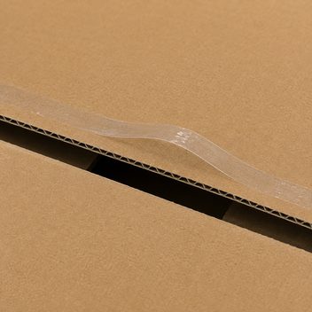 A close view along the centre seam of a plain kraft shipping carton where a single strip of clear tape, far narrower than the seam it is closing, has released over the middle of its run and lifted clear of the surface; beneath it the top flaps have sprung apart into an open gap with the corrugated flute structure showing along the exposed edges, while the tape stays stuck down at either end.