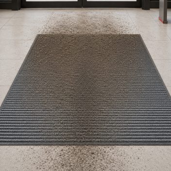 A ribbed entrance mat inside a set of glazed doors, its far edge packed and clogged with grey-brown grit where shoes first land and fading gradually toward the interior, a darker traffic lane worn down the middle; the same soil lies tracked on the pale tile beyond the entry edge and again on the tile past the near end, carried straight across the mat and into the building.
