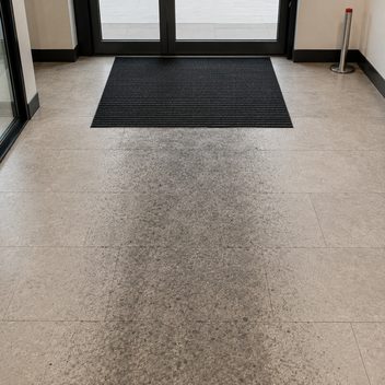 An entry mat just inside a pair of glazed doors, only two or three paces deep, with a broad band of dark soiling worn into the matte tile floor beginning exactly at the mat's inner edge and tracking away into the building; the floor to either side of the walking path is clean and pale.