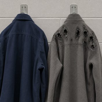 Two work garments hung backs-out on hooks against a painted concrete wall; the navy cotton work shirt on the left is intact and sound, while the grey synthetic fleece beside it has melted across the back of the shoulder and upper sleeve into a cluster of holes with hard glossy shrivelled rims, the pile around them fused smooth against the soft matte fleece below.