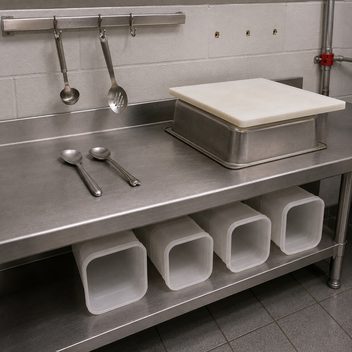 A prep station carrying four different people's workarounds at once: a wall rail with five hooks but only two in use, both holding utensils too small to seat and hanging askew; three bare screw holes beside it where a second rail was removed; an upturned food pan propping a cutting board off the counter; and four containers stored on their sides beneath because the shelf clearance is too low to stand them up.
