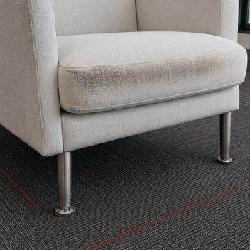 A pale upholstered lounge chair cropped close, a band of dark ingrained soiling worn into the front edge of the seat cushion where people push themselves up, the same cushion clean and pale everywhere else; the glide on the near front leg is chewed and scuffed raw while the far leg's glide is intact.