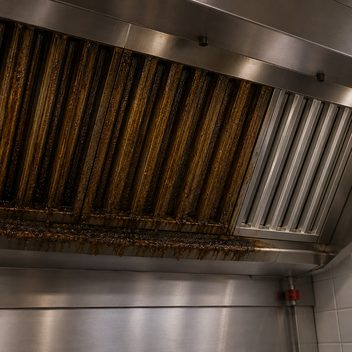 The underside of a commercial kitchen exhaust hood seen from below, its baffle filters packed and glazed with thick amber-brown grease that has bridged the slots closed and run down to harden in drips along the lower lip, the collection channel overrun; at one end of the same bank a single recently cleaned filter stands bright bare stainless with its channels open and clear.