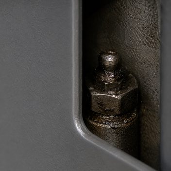 A grease fitting set deep in a narrow gap behind the edge of a machine guard panel, the fitting crusted and darkened with old grease while the painted panel around it is clean.