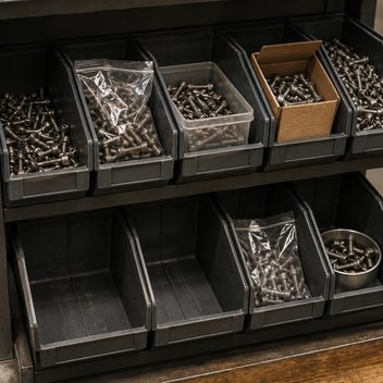 Two shelves of parts-crib bins holding the same socket-head fastener in five different forms — loose, bagged, in a plastic tub, in a cardboard box and in a metal tin — with several bins empty.