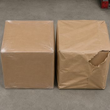 Two identical kraft shipping cartons standing side by side, the one on the left wrapped tight and square in clear plastic film, the one on the right wrapped in brown paper that hangs slack and bagging at the corners and has torn open along a ragged fibrous line, exposing the intact corrugated of the box underneath.