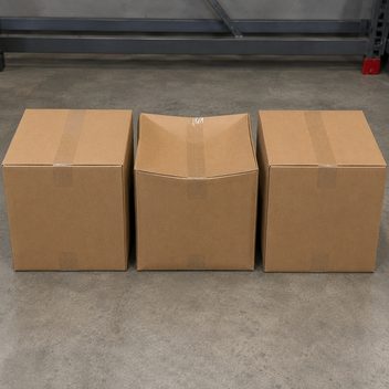 Three identical plain kraft shipping cartons standing sealed in a row on a concrete floor, the ones at either end square and full with flat firm top faces, while the middle carton of the same size has sagged inward across its top and drawn in at the sides where the tape holds a lid over mostly empty air.