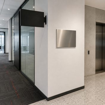 A corridor beside a glazed meeting room, with a blank matte black projecting blade sign mounted above the glass and a blank brushed stainless plaque fixed flat to the wall a short distance away, the two signs different sizes, finishes and mounting planes with nothing written on either.