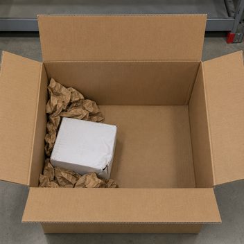 A large plain kraft shipping carton standing open with a single small white carton sitting in one corner, a thin wad of crumpled kraft paper heaped around it and more than half the box interior left as bare empty space; the white carton has one corner crushed inward and its flap split, the face beside it buckled and scuffed grey where it slid against the corrugated wall, while the shipping carton itself is clean and square.