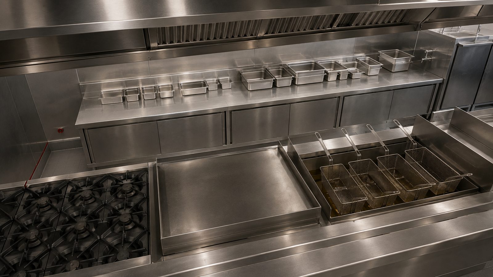 A commercial kitchen hot line seen from above and behind, the open burner grates of a range, a flat griddle and a bank of fryers with wire baskets on their rails laid out as horizontal work surfaces, a stainless prep counter beyond them lined with clean unmarked pans, and the underside of the exhaust hood crossing above.